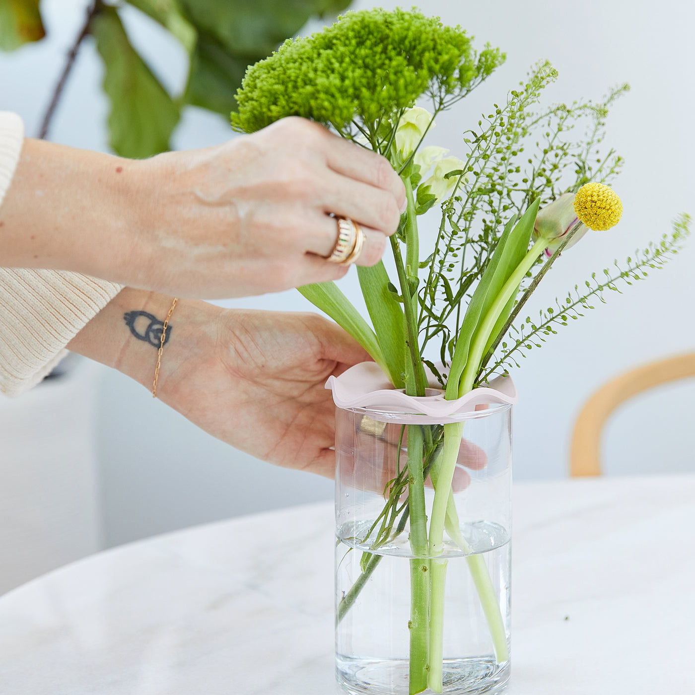 A hand arranges vibrant green and yellow flowers in a clear glass vase on a marble surface, enhancing indoor decor.