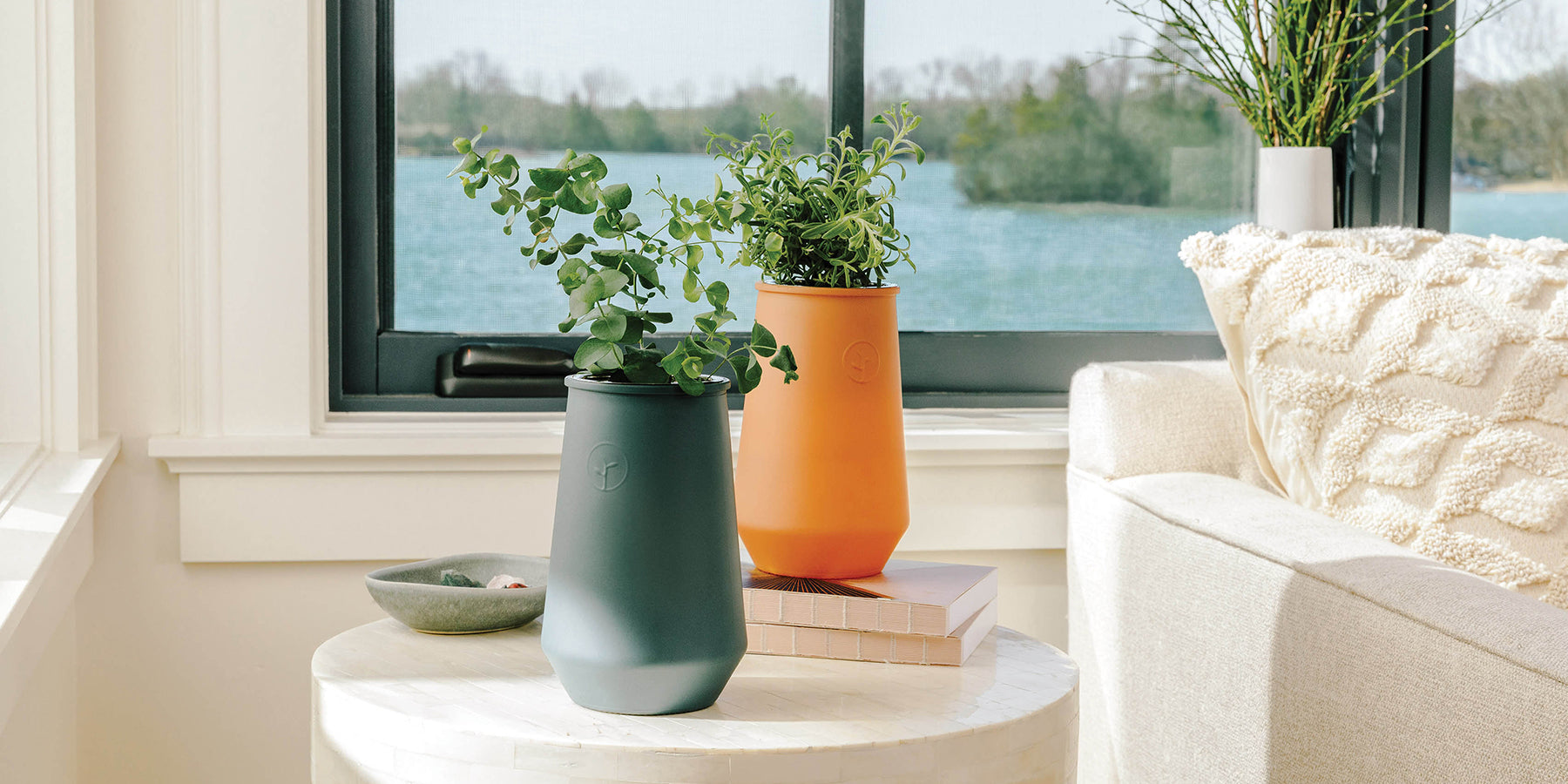 Two stylish hydroponic pots in teal and orange, featuring fresh herbs, set on a white table near a sunny window.