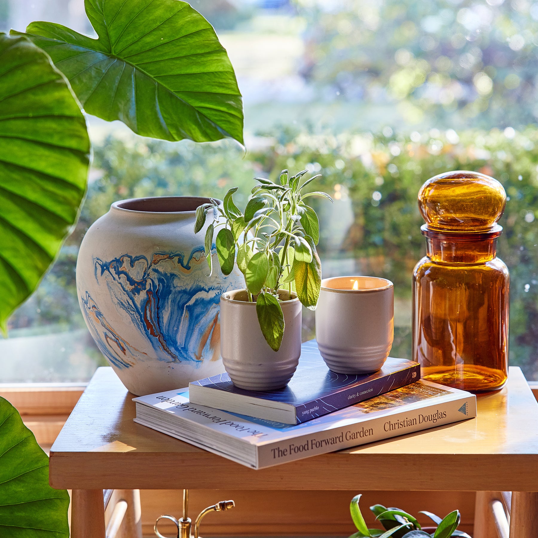 A stylish 2-in-1 candle and planter set featuring a small plant, stacked books, and decorative glassware on a wooden table.
