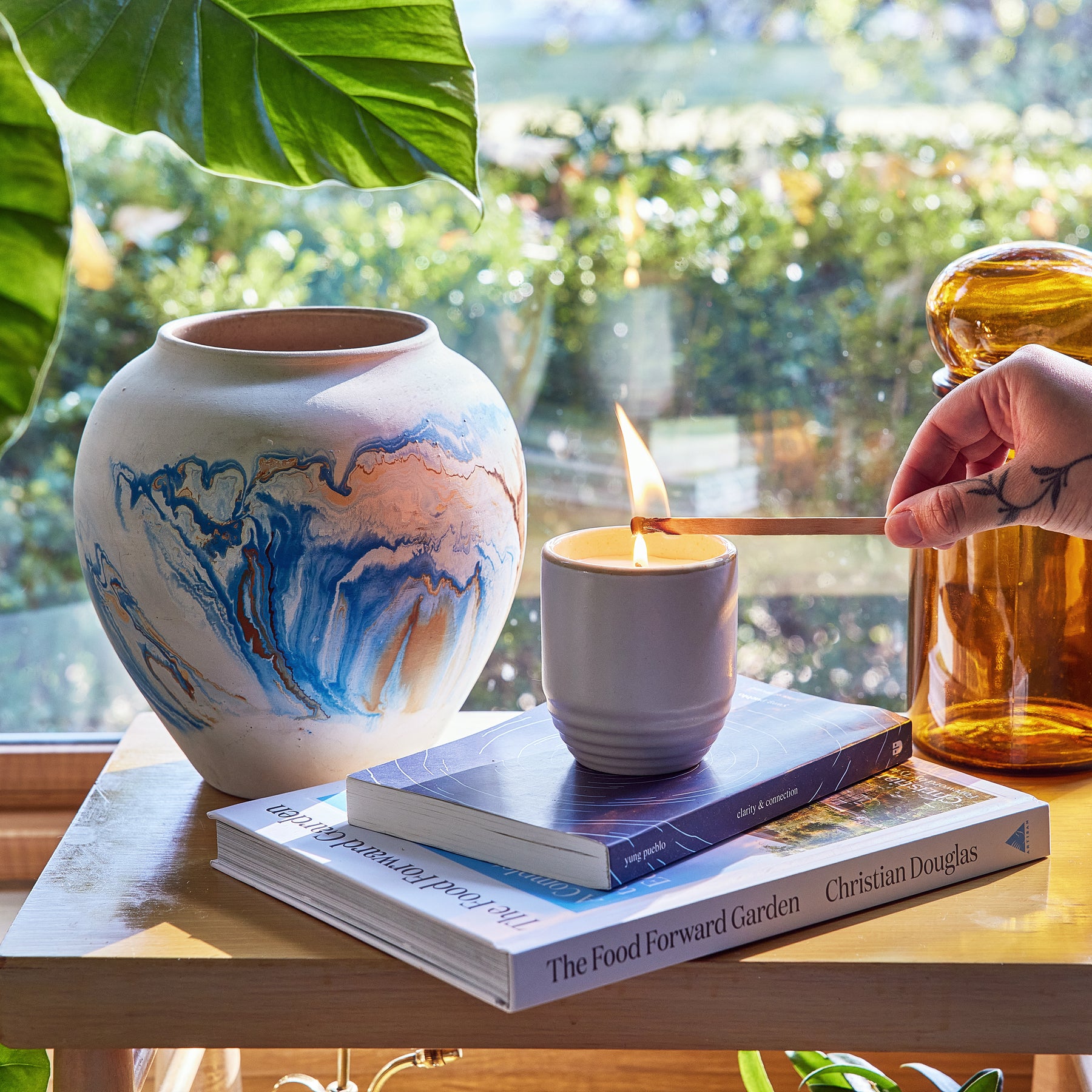 A hand lights a candle in a ceramic planter, surrounded by books and greenery, showcasing a cozy indoor garden vibe.