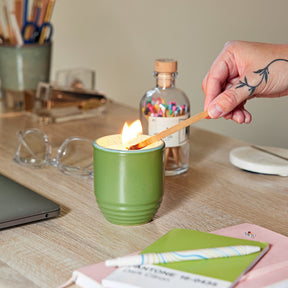 A hand lights a green candle in a textured planter on a wooden desk, surrounded by office supplies and a laptop.