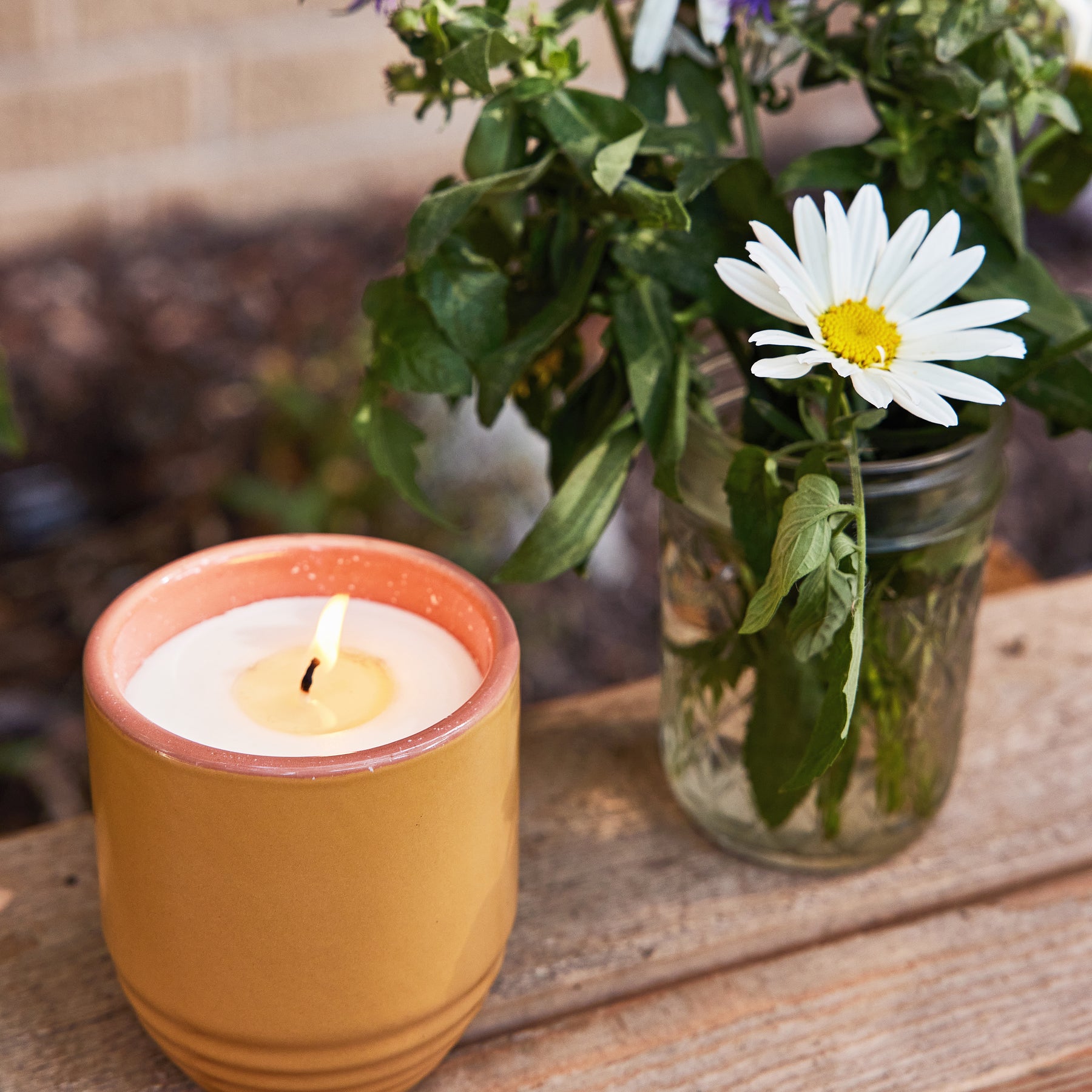 A yellow candle planter with a lit candle beside a glass jar holding a daisy and green foliage on a wooden surface.