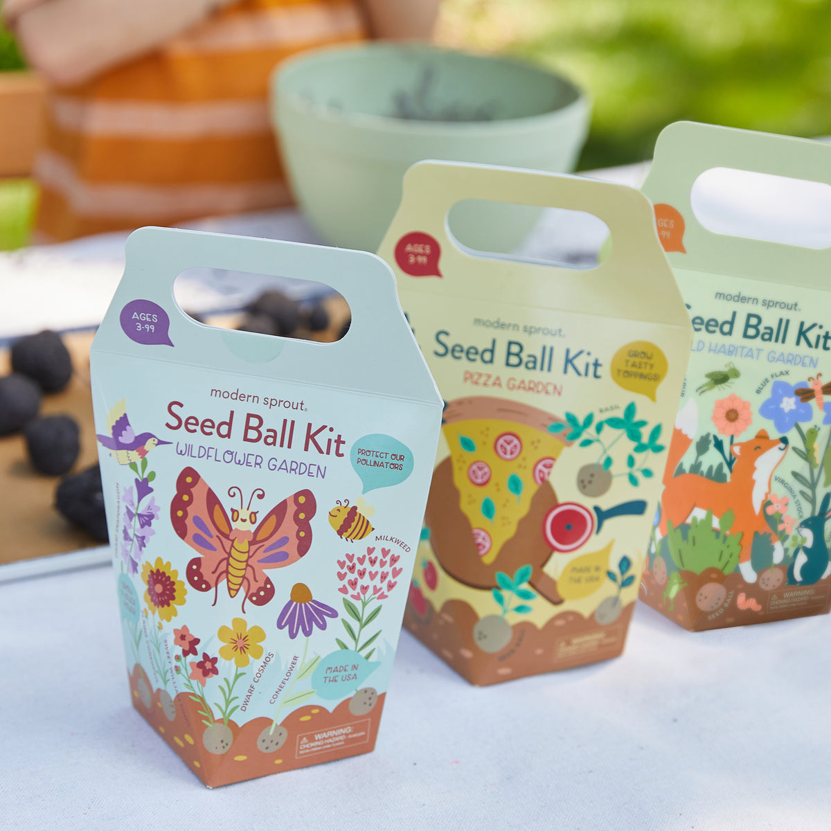 Seed Ball Kits displayed outdoors, featuring Wildflower, Pizza Garden, and Habitat Garden options, promoting gardening fun.