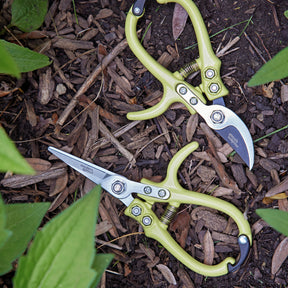 Gardening shears and pruners with green handles resting on mulch among plants, showcasing sharp stainless steel blades.