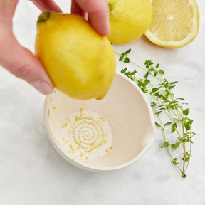 A hand holds a lemon above a ceramic dish with zest inside, surrounded by fresh lemons and thyme sprigs.