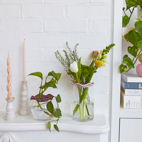 Two Botanical Keepers holding fresh flowers in vases on a white shelf, alongside decorative candles and plants.