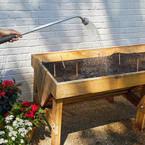 A hand waters a wooden planter filled with soil and seed pops, surrounded by blooming flowers against a white wall.