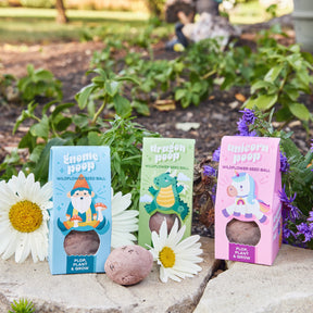 Three colorful seed ball boxes labeled Gnome Poop, Dragon Poop, and Unicorn Poop, surrounded by flowers and greenery.