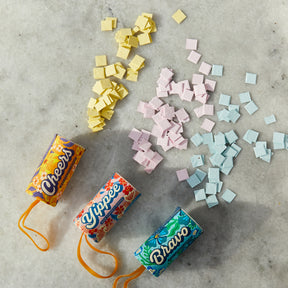 Colorful seed confetti poppers in vibrant packaging, surrounded by pastel seed confetti pieces on a marble surface.