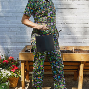 A person in a colorful floral outfit holds a sleek black watering can next to a wooden planter filled with soil.