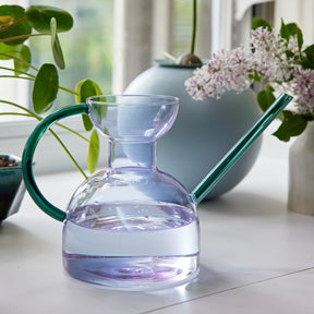 Elegant glass watering can with a curved handle and spout, filled with water, surrounded by indoor plants.