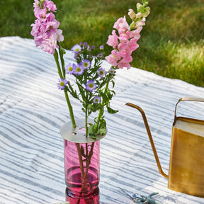 Brass flower frog atop a pink vase holding pink and purple flowers on a striped fabric outdoors.