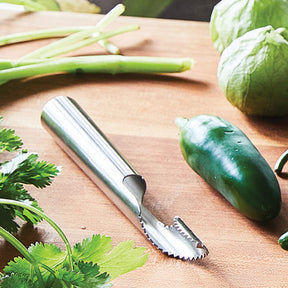 Stainless steel pepper de-seeder tool on a wooden surface surrounded by fresh vegetables and herbs.
