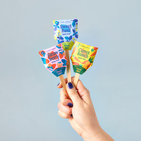 Hand holding three colorful Seed Pops for gardening: Sweet Basil, Strawberry, and Carrot Bunch, against a light background.