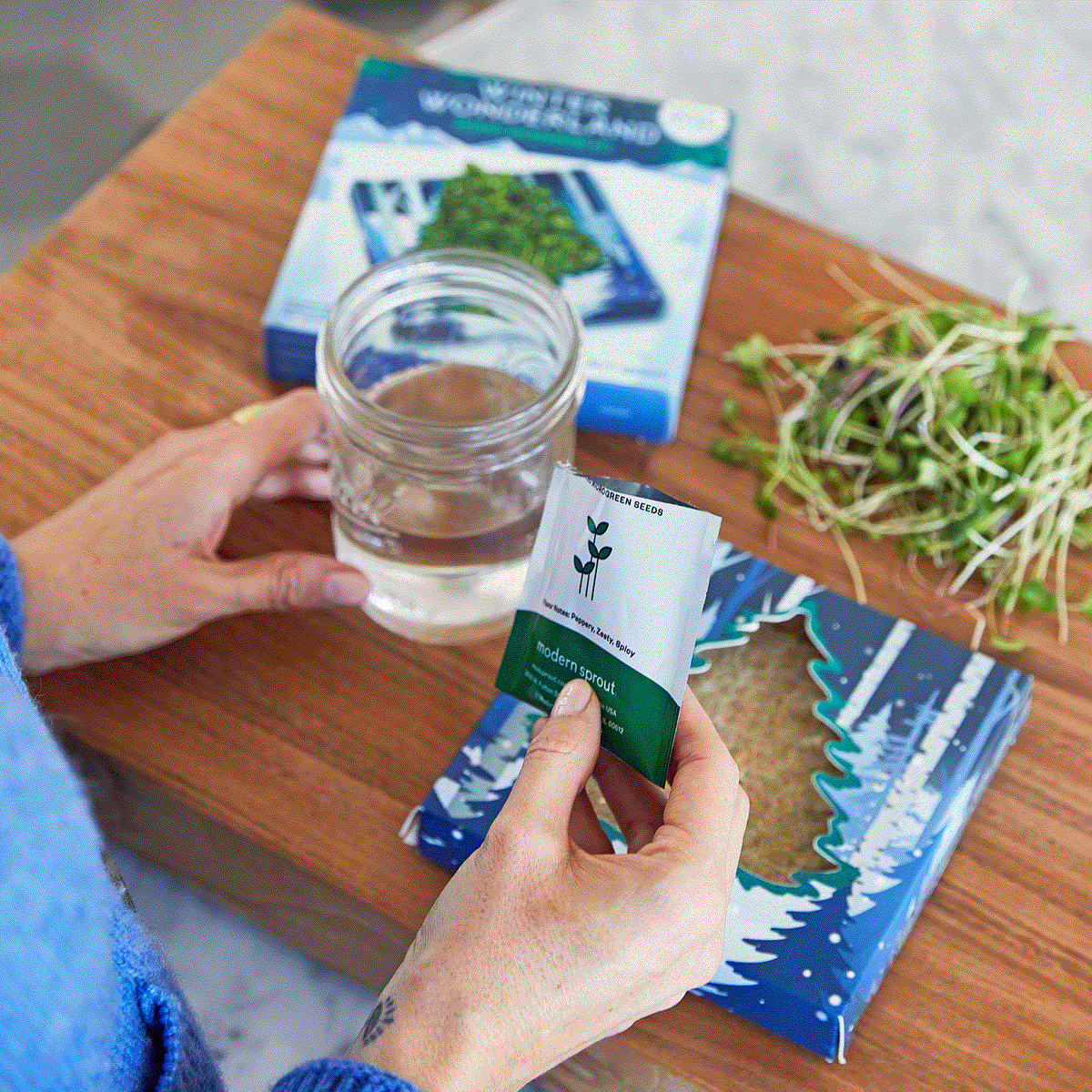 A person holds a seed packet above a glass of water, with a hydroponic growing kit box in the background.