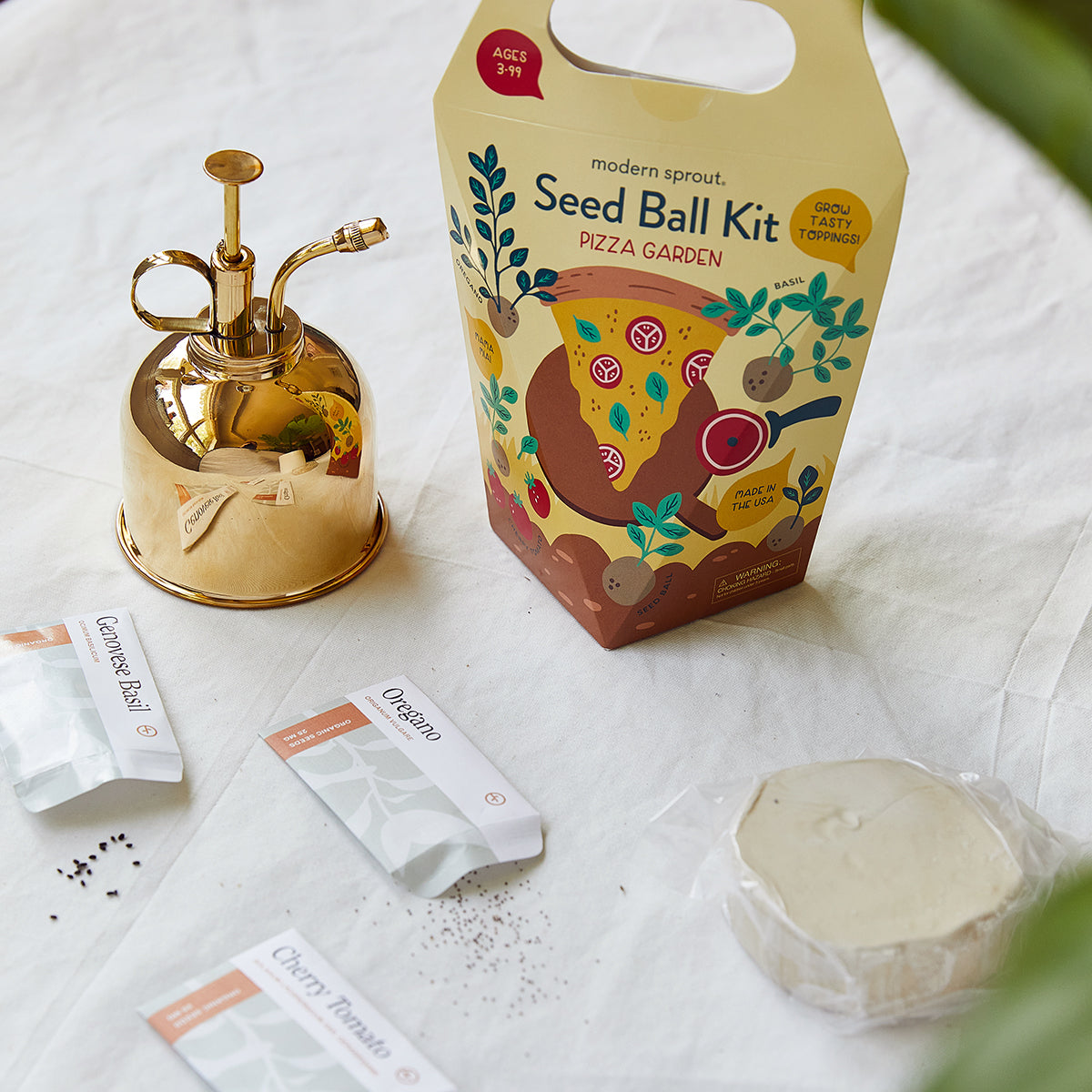 Gold watering can next to a "Pizza Garden" seed ball kit and seed packets on a white cloth.