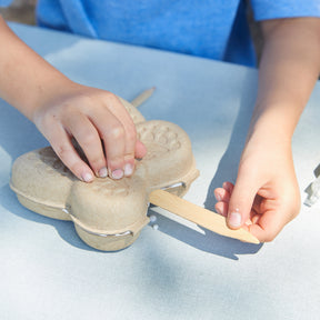 Child's hands opening a Curious Critters Garden Kit, revealing planting materials and tools on a table outdoors.