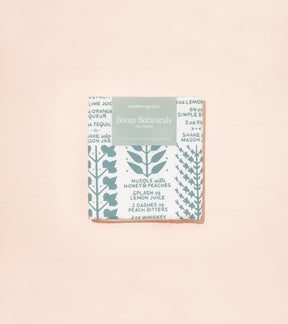Boozy Botanicals tea towel featuring a botanical print with recipe text in blue and white, packaged neatly.